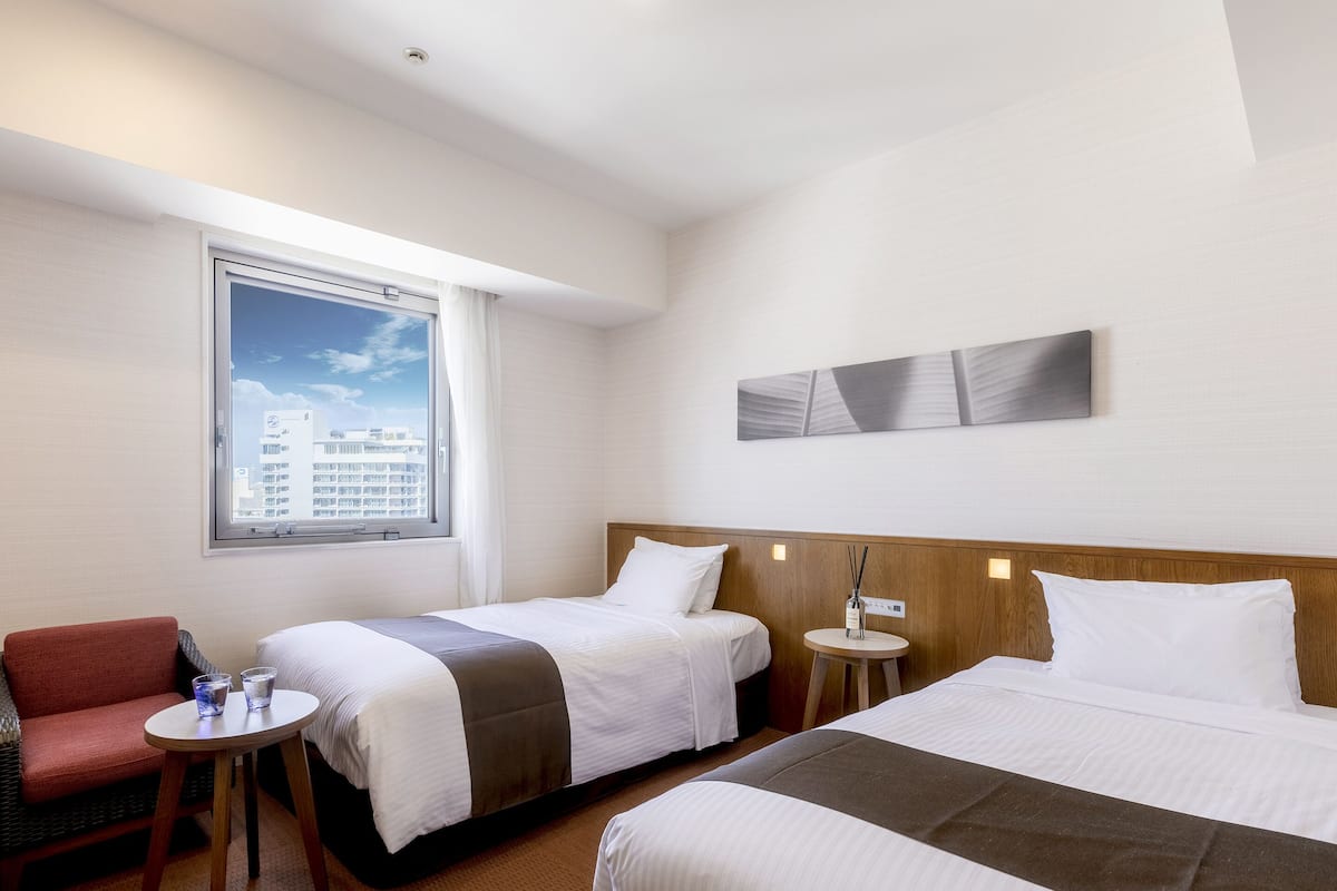 Hotellihuone, jossa on kaksi sänkyä, pieni pöytä lasineen, punainen tuoli ja ikkuna, josta avautuu näkymä kaupunkiin.