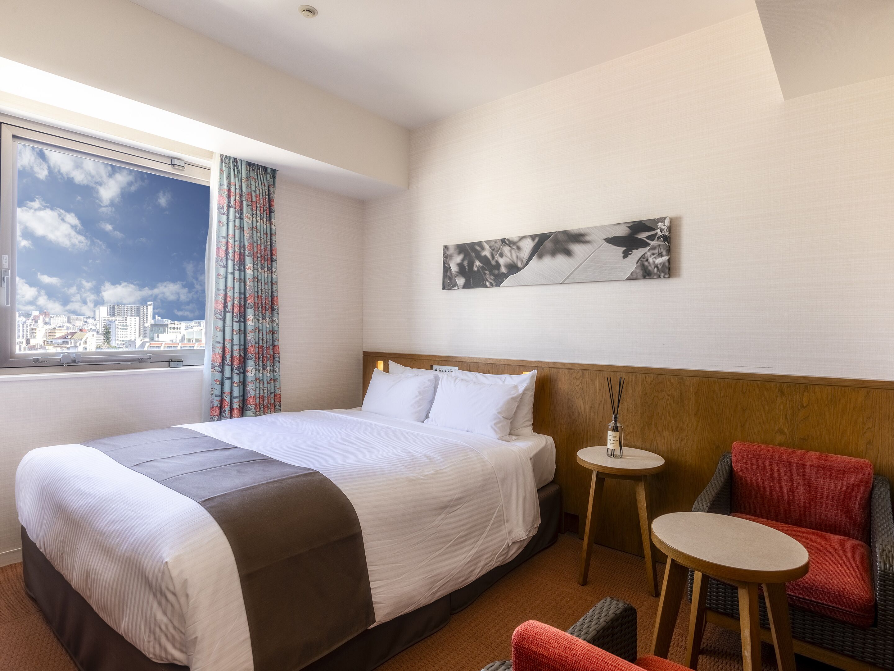 A hotel room with a bed, a small table, a chair, and a view of the city through a window.