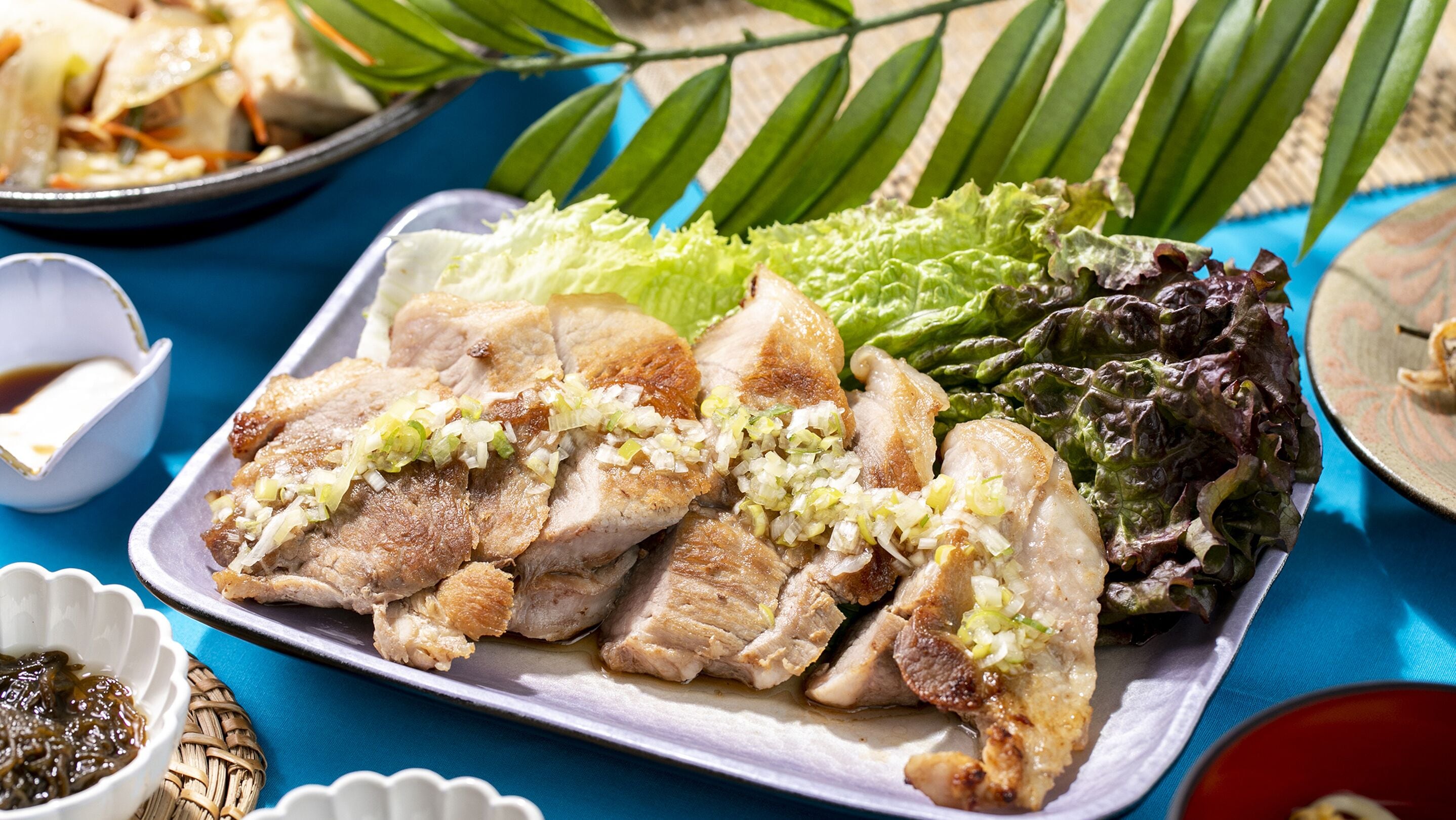 A plate of sliced meat garnished with chopped onions, accompanied by mixed green and purple lettuce leaves, on a blue tablecloth.