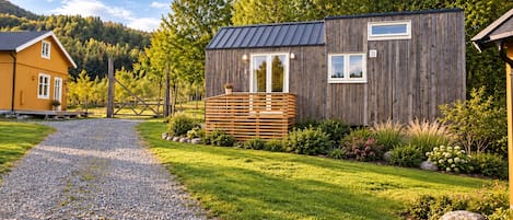 A wooden cabin with a gravel path leading to it, surrounded by greenery and a mountain backdrop.