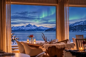 Un comedor con una mesa preparada para dos personas, vista a montañas nevadas y un lago.