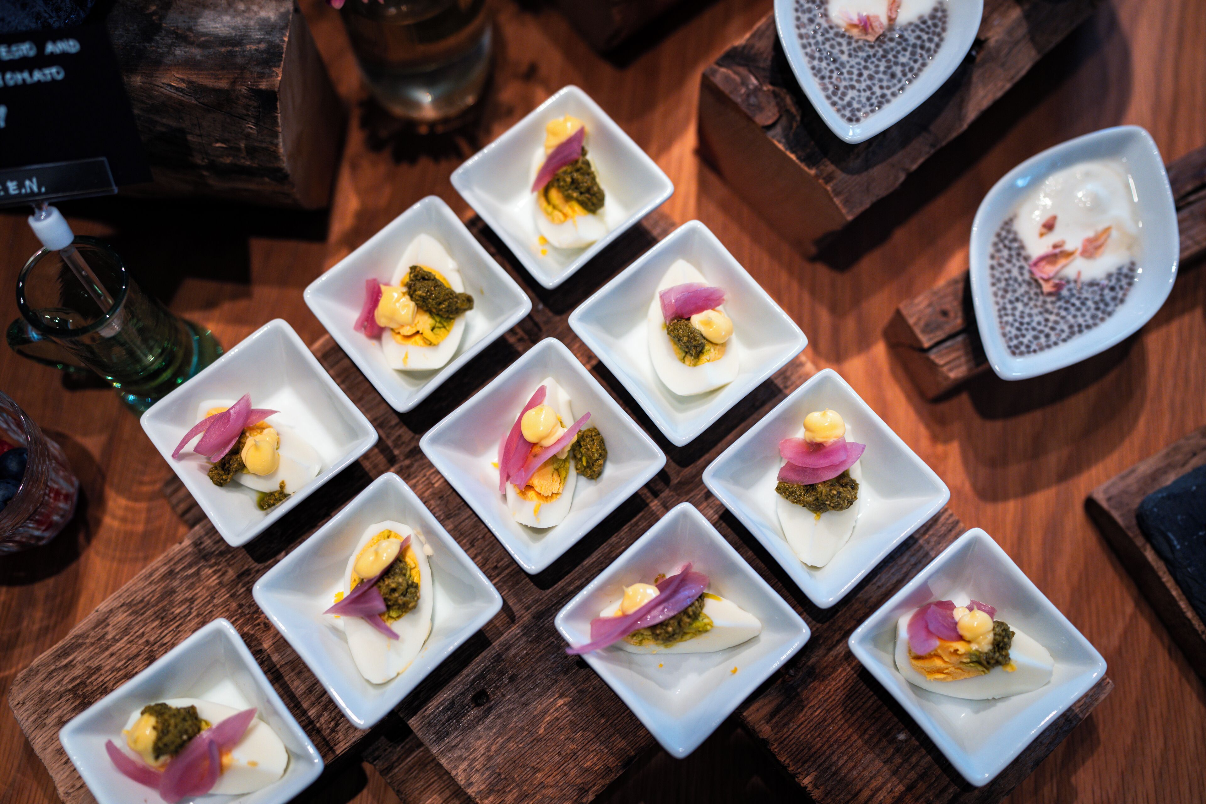 A wooden table with multiple square white dishes containing food items, likely appetizers, arranged neatly.