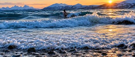 A beach with waves, a person in the water, and a sunset with mountains in the background.