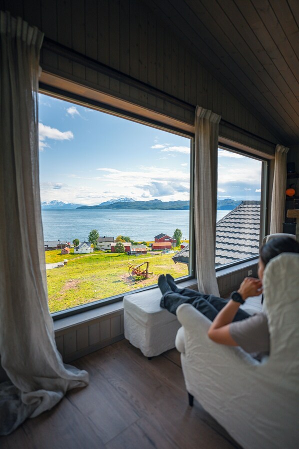 A room with a large window overlooking a lake and mountains. There is a person sitting on a white chair with a view of the landscape. - Yggdrasil Farmhotel Retreat, Spa & Yoga (Tromsø)