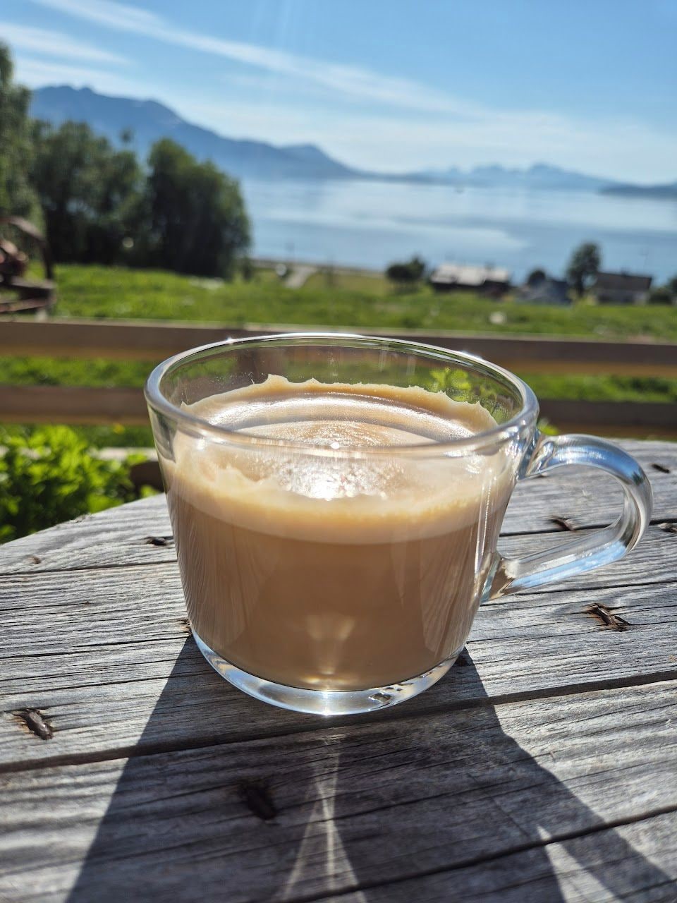 Una taza de vidrio con café sobre una mesa de madera, con una vista panorámica de montañas y vegetación al fondo.