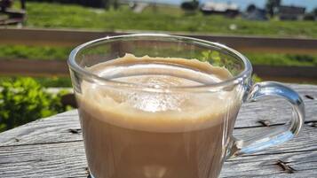 Una taza de vidrio con café sobre una mesa de madera, con una vista panorámica de montañas y vegetación al fondo.