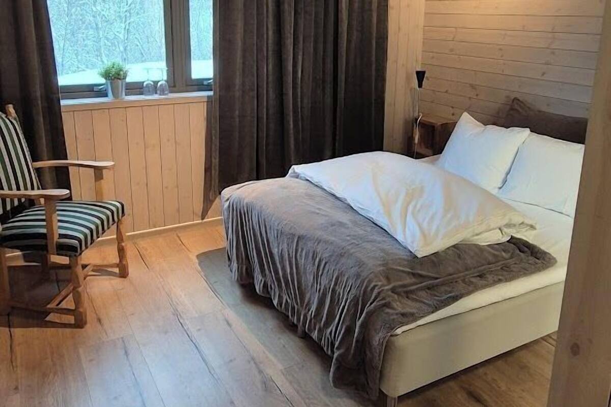 Ein Schlafzimmer mit Holzwänden, einem Bett, einem Sessel, einem Fenster mit Vorhängen und einer Heizung.