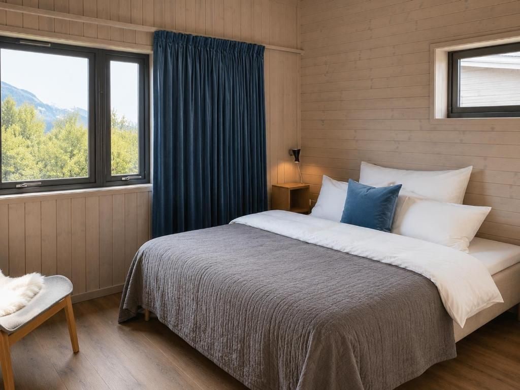 Un dormitorio con cama, una silla, un ventanal con cortinas y una mesita de noche con lámpara.