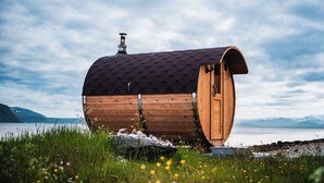 A wooden barrel-shaped structure with a chimney, situated by a lake with mountains in the background. - Yggdrasil Farmhotel Retreat, Spa & Yoga (Tromsø)