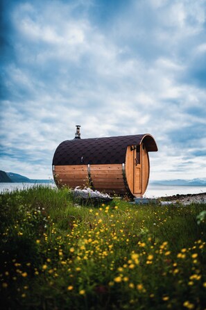 A wooden barrel-shaped structure with a chimney, situated by a lake with mountains in the background. - Yggdrasil Farmhotel Retreat, Spa & Yoga (Tromsø)