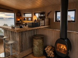 A rustic cabin interior with a wooden bar, a wood-burning stove, and a view of the water. - Yggdrasil Farmhotel Retreat, Spa & Yoga (Tromsø)