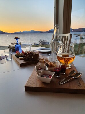 A table set with various food items and drinks, a candle, and a view of a lake at sunset. - Yggdrasil Farmhotel Retreat, Spa & Yoga (Tromsø)