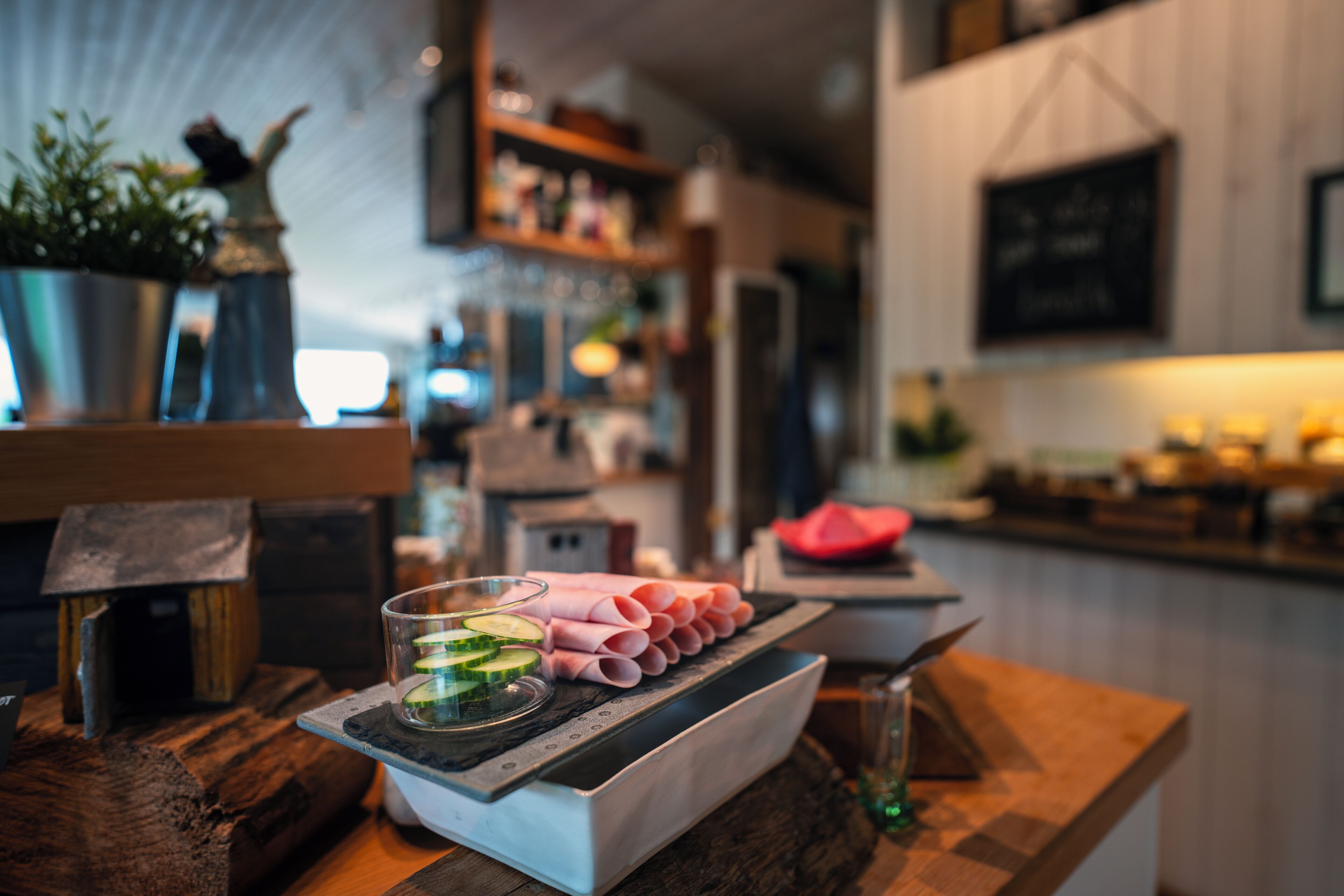 Interior de restaurante con una mesa de madera que presenta pepinos en rodajas y jamón, un vaso de agua y un comedero decorativo para pájaros.