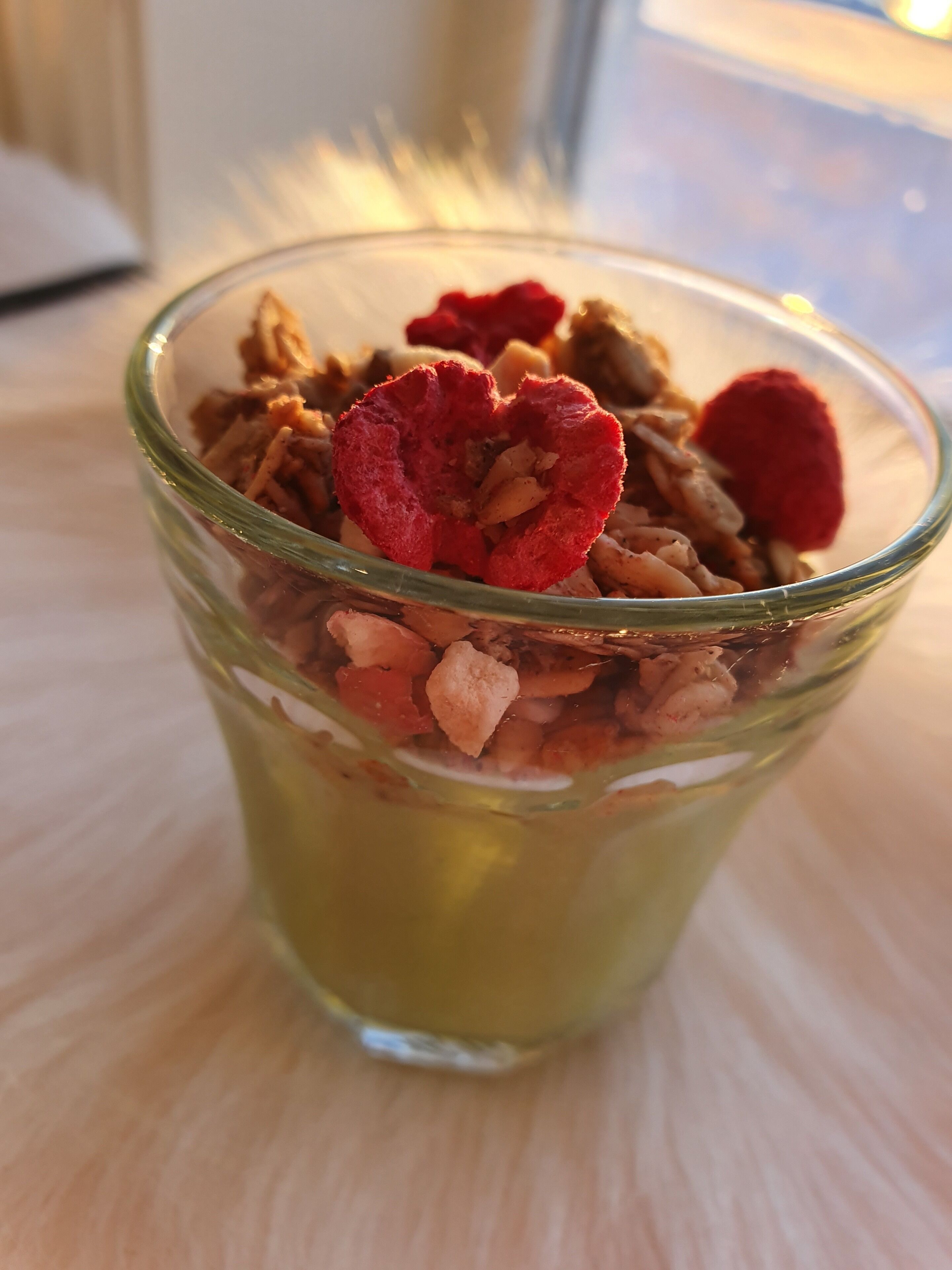 A glass bowl containing granola with red fruit pieces on a light-colored surface.