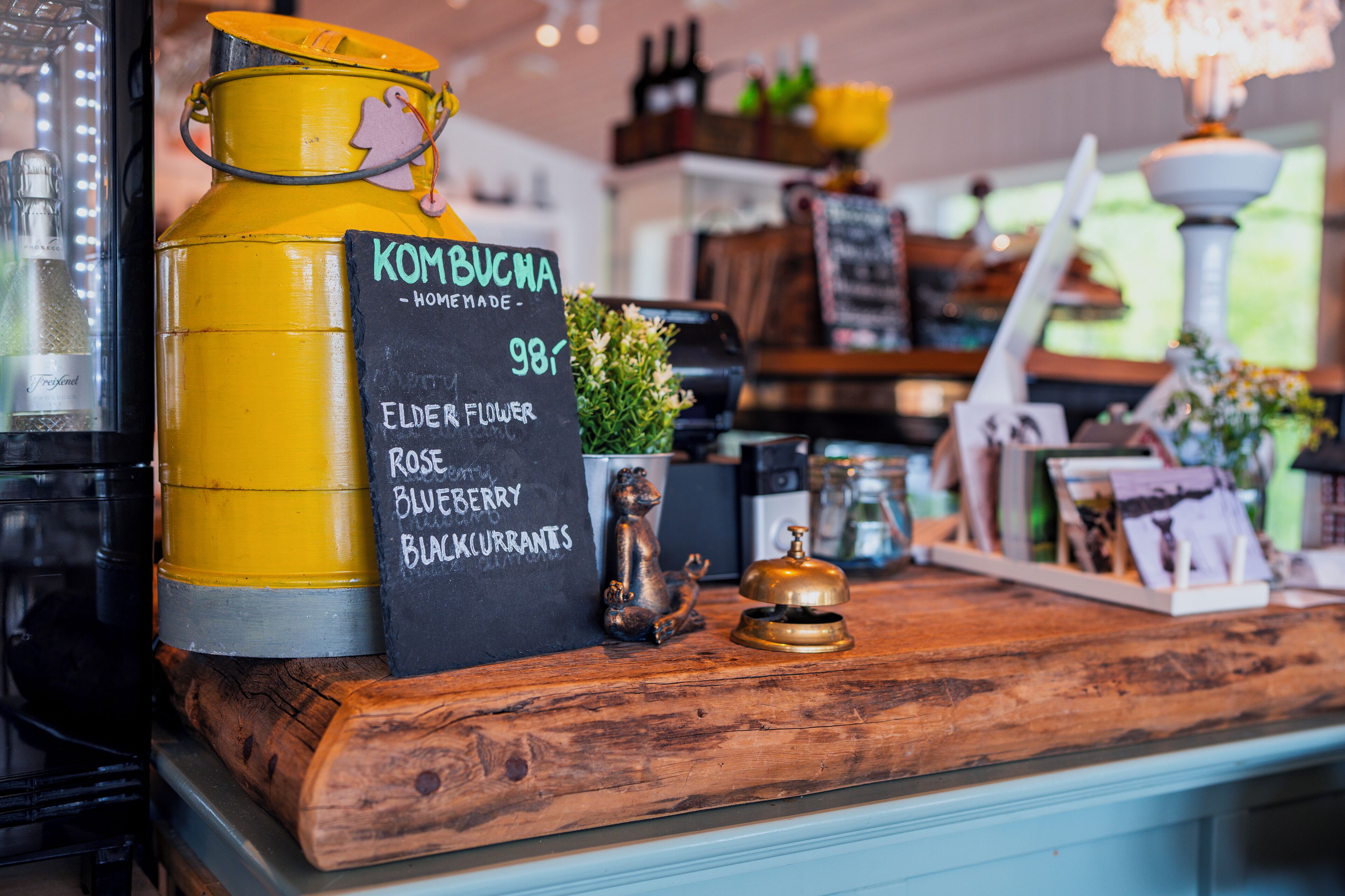 Een houten toonbank met een gele container met de tekst 'KOMBUCHA' en een bord waarop 'elderbloesem, roos, bosbessen, zwarte bessen' staat.