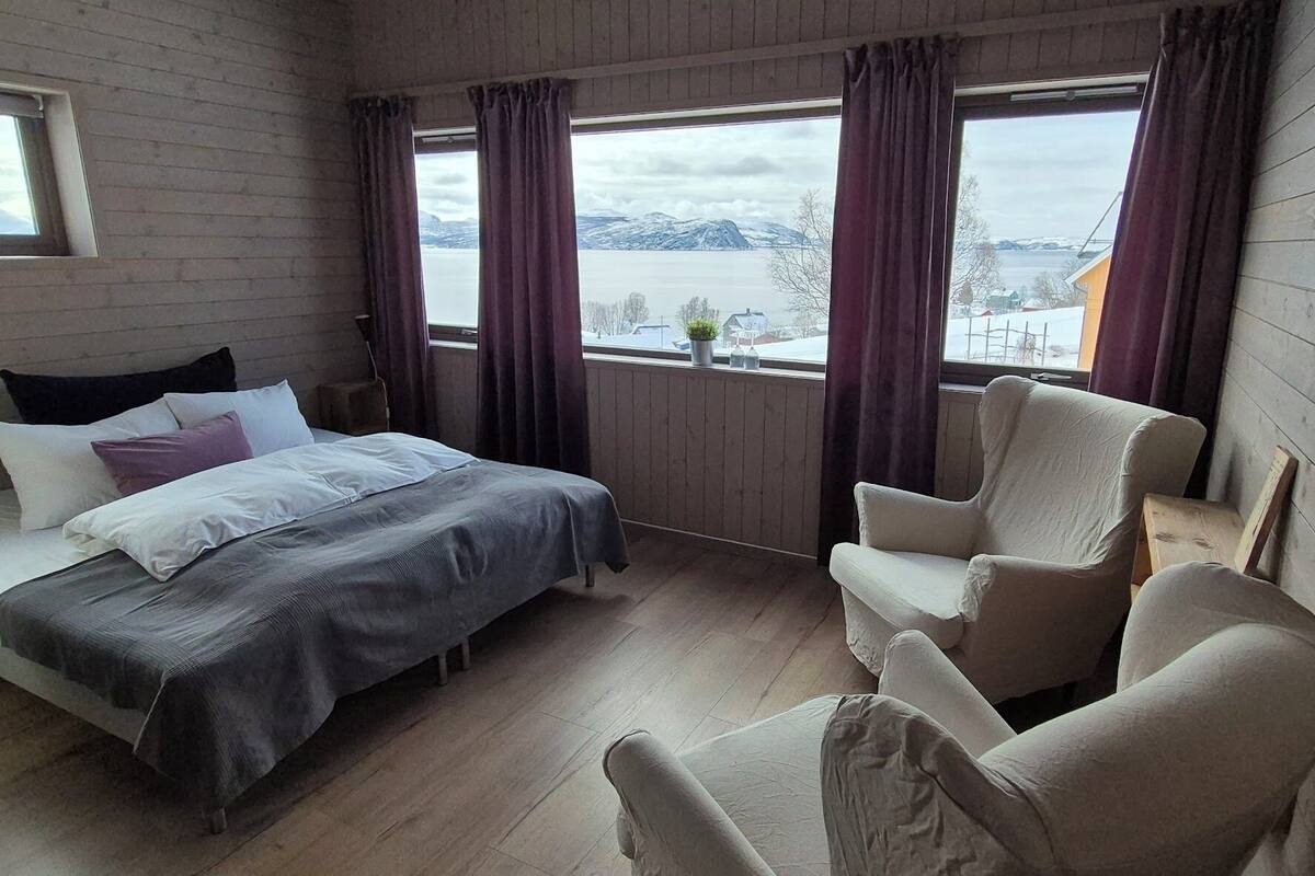 Ein Schlafzimmer mit einem Bett, zwei Stühlen, einem Fenster mit Vorhängen und Blick auf einen See und Berge.