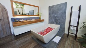 Un baño moderno con una bañera independiente llena de pétalos de rosa, un tocador de madera, un espejo grande y un perchero para toallas.