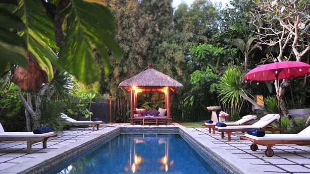 Un área de piscina con sillones, un pabellón de techo de paja y abundante vegetación.