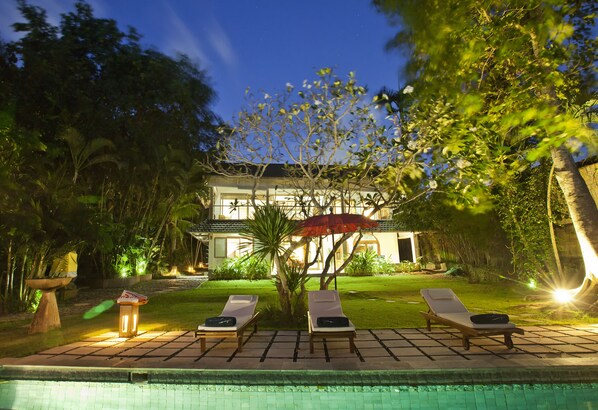 A poolside area with lounge chairs and a red umbrella, surrounded by lush greenery and a house in the background.