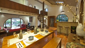 Un amplio espacio habitable con una larga mesa de madera dispuesta para comer, una escalera y grandes ventanales que ofrecen vistas al exterior.