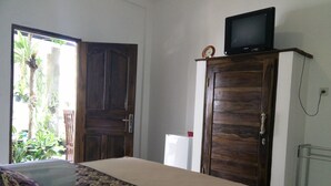 A room with a bed, a TV on a wooden cabinet, a white refrigerator, and a door leading to a balcony with plants. - Double N Guesthouse (Denpasar)