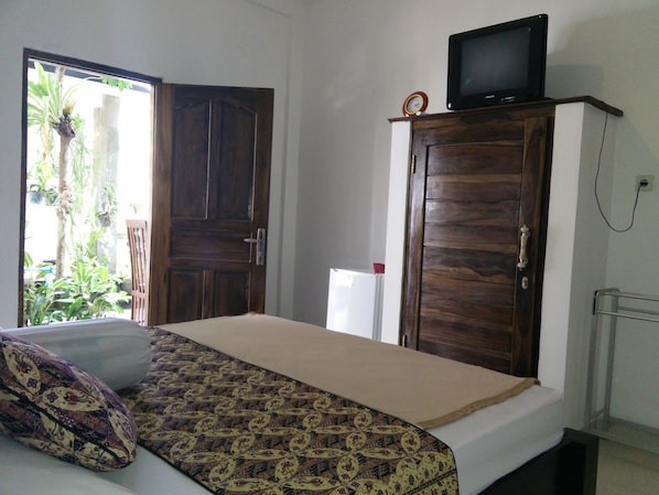 A room with a bed, a TV on a wooden cabinet, a white refrigerator, and a door leading to a balcony with plants. - Double N Guesthouse (Denpasar)