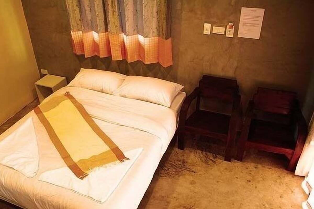 A single bed with a white comforter and a beige throw blanket, a nightstand, a wooden chair, and a small table with a lamp.