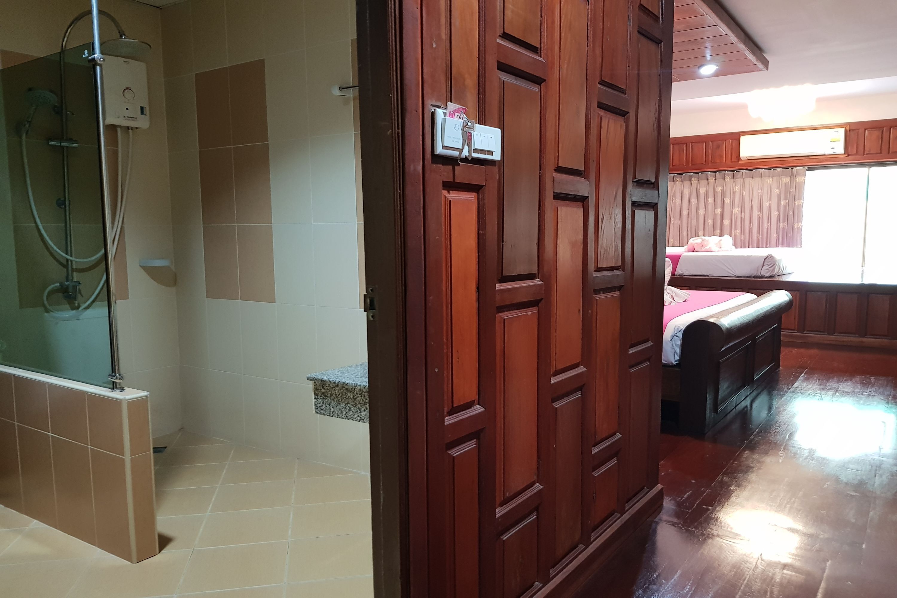 Casa de banho com chuveiro, sanita e lavatório. Uma porta de madeira dá acesso a um quarto com uma cama e uma janela.
