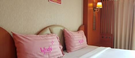 Una habitación de hotel con una cama, dos almohadas rosas con texto, una pared con un cartel de piña y una ventana con cortinas.