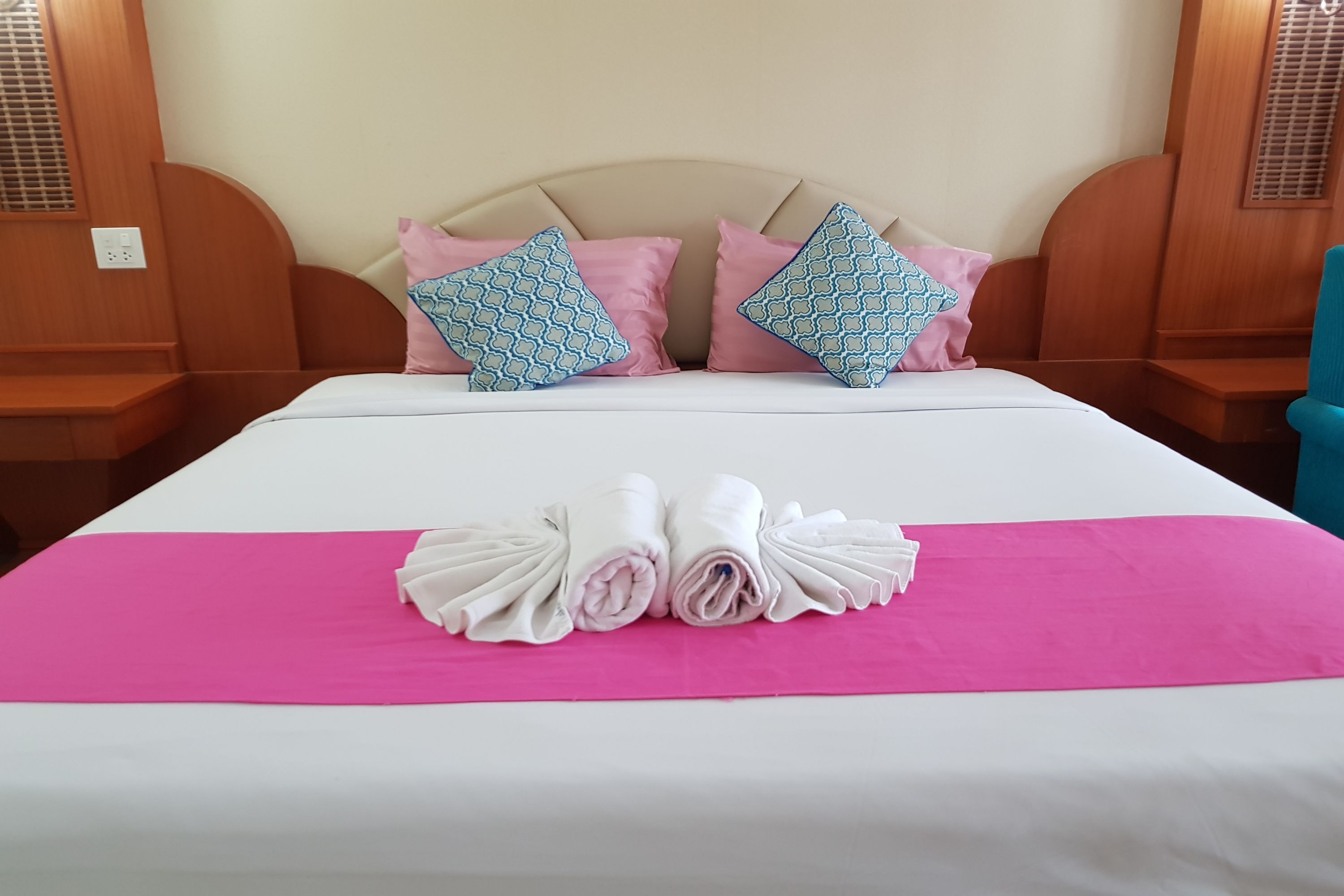A neatly made bed with pink and blue patterned pillows, a pink bedspread, and rolled towels on a white bed.