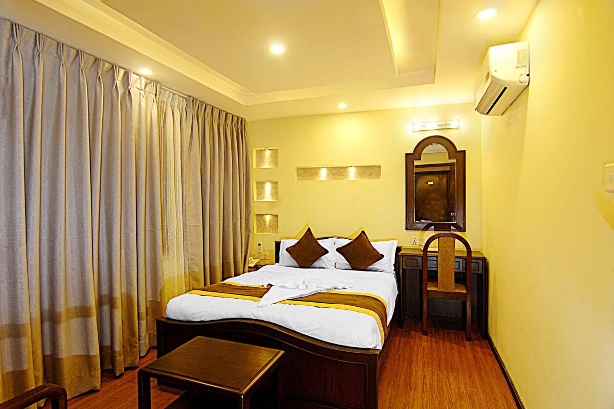 A hotel room with a large bed, bedside tables, a chair, a desk, and an air conditioning unit.