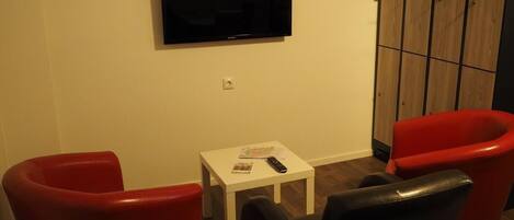 A room with a flat-screen TV mounted on the wall, a small white table with a remote control, and two red chairs.