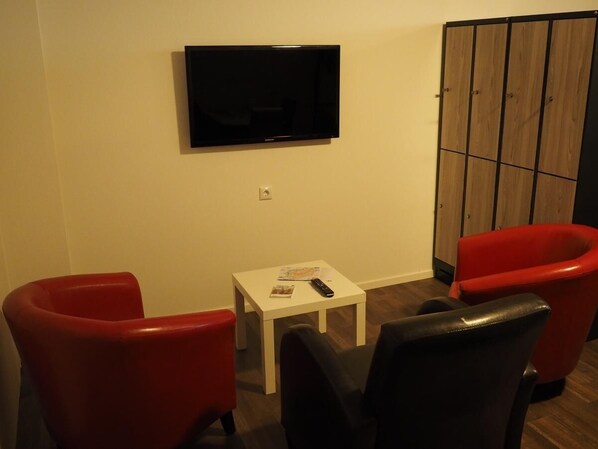 Habitación con una televisión de pantalla plana montada en la pared, una mesita blanca con un control remoto y dos sillas rojas.