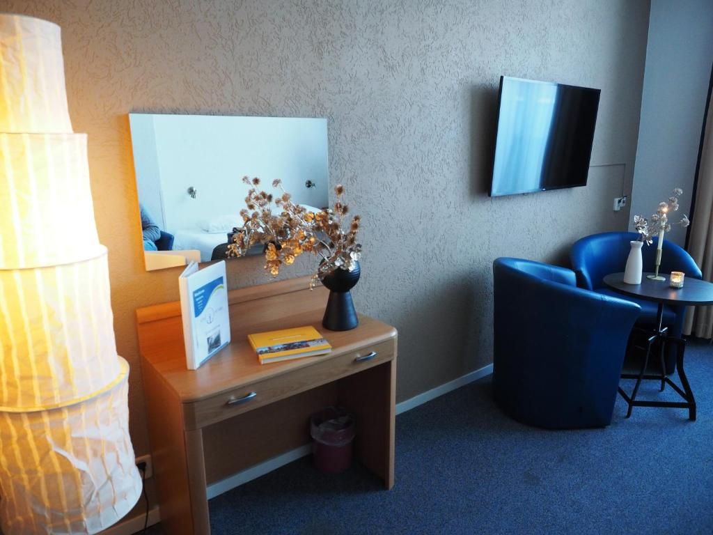 A hotel room with a desk, a lamp, a vase with dried flowers, a blue chair, a TV mounted on the wall, and a small table with a candle.