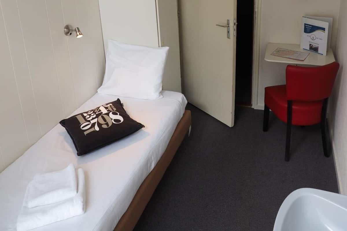 A small, neatly arranged room with a bed, a desk, a chair, and a bathroom.