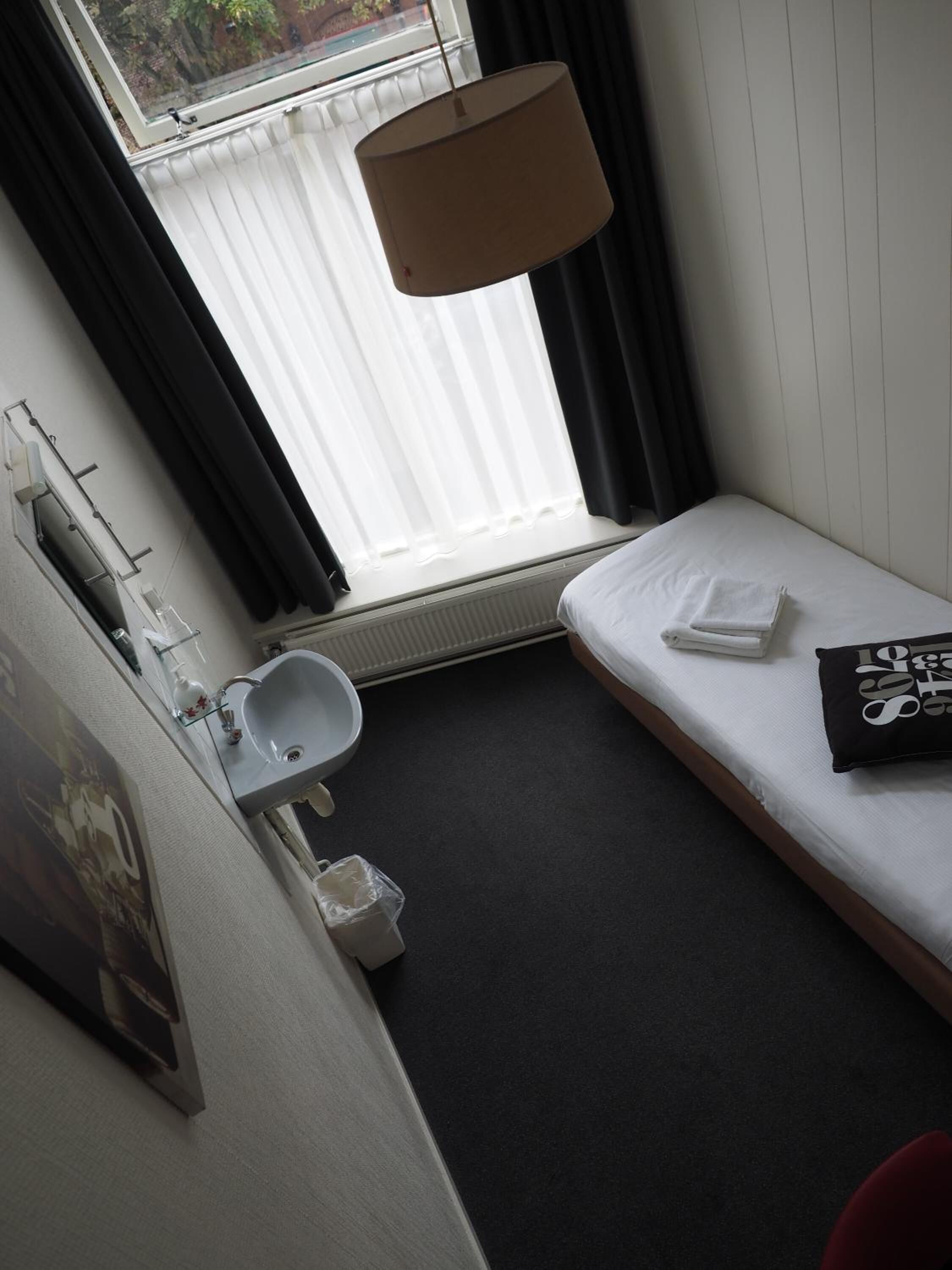 A small, clean room with a single bed, a bedside table, a window with curtains, a hanging lamp, a sink, and a towel.