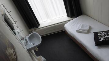 A small, clean room with a single bed, a bedside table, a window with curtains, a hanging lamp, a sink, and a towel.