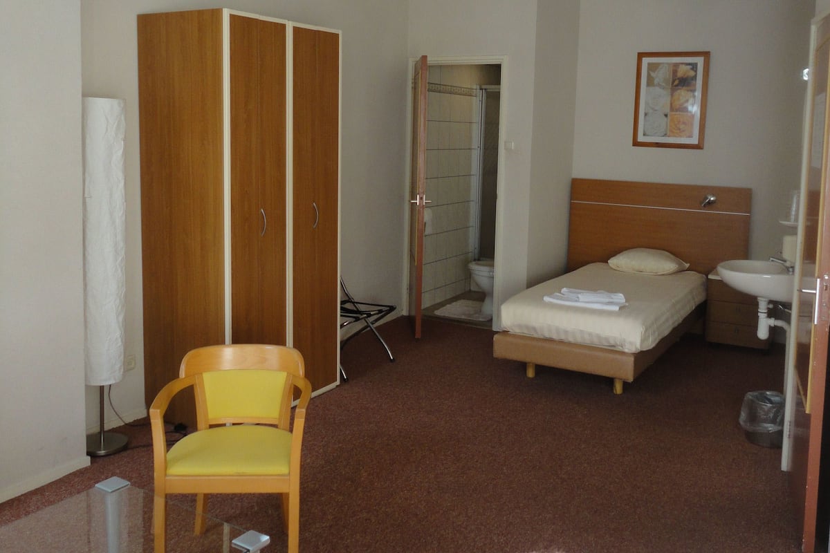 A small, compact hotel room with a single bed, a bedside table, a chair, a wardrobe, and a bathroom with a toilet and sink.