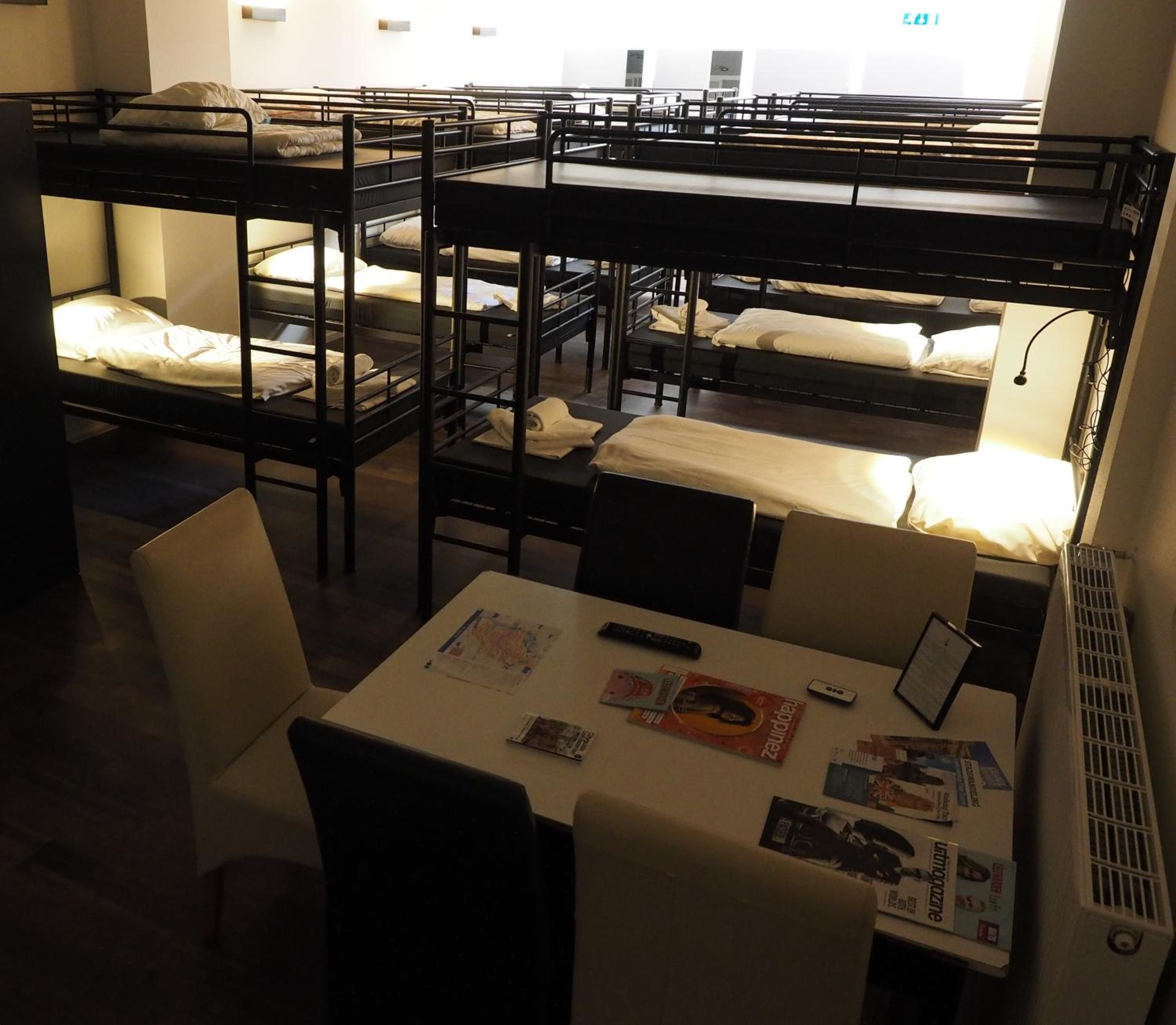 Un dormitorio con literas, una mesa con revistas y un ventanal con letrero de salida.