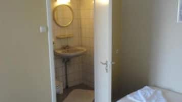 Una habitación de hotel pequeña y sencilla con una cama, una mesita de noche, un baño pequeño con lavamanos y espejo, y una puerta que da al baño.
