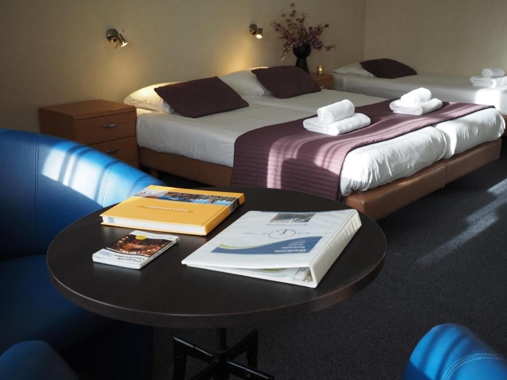 Una habitación de hotel con dos camas, una mesa redonda con libros, una silla azul y una mesita de noche con una lámpara.