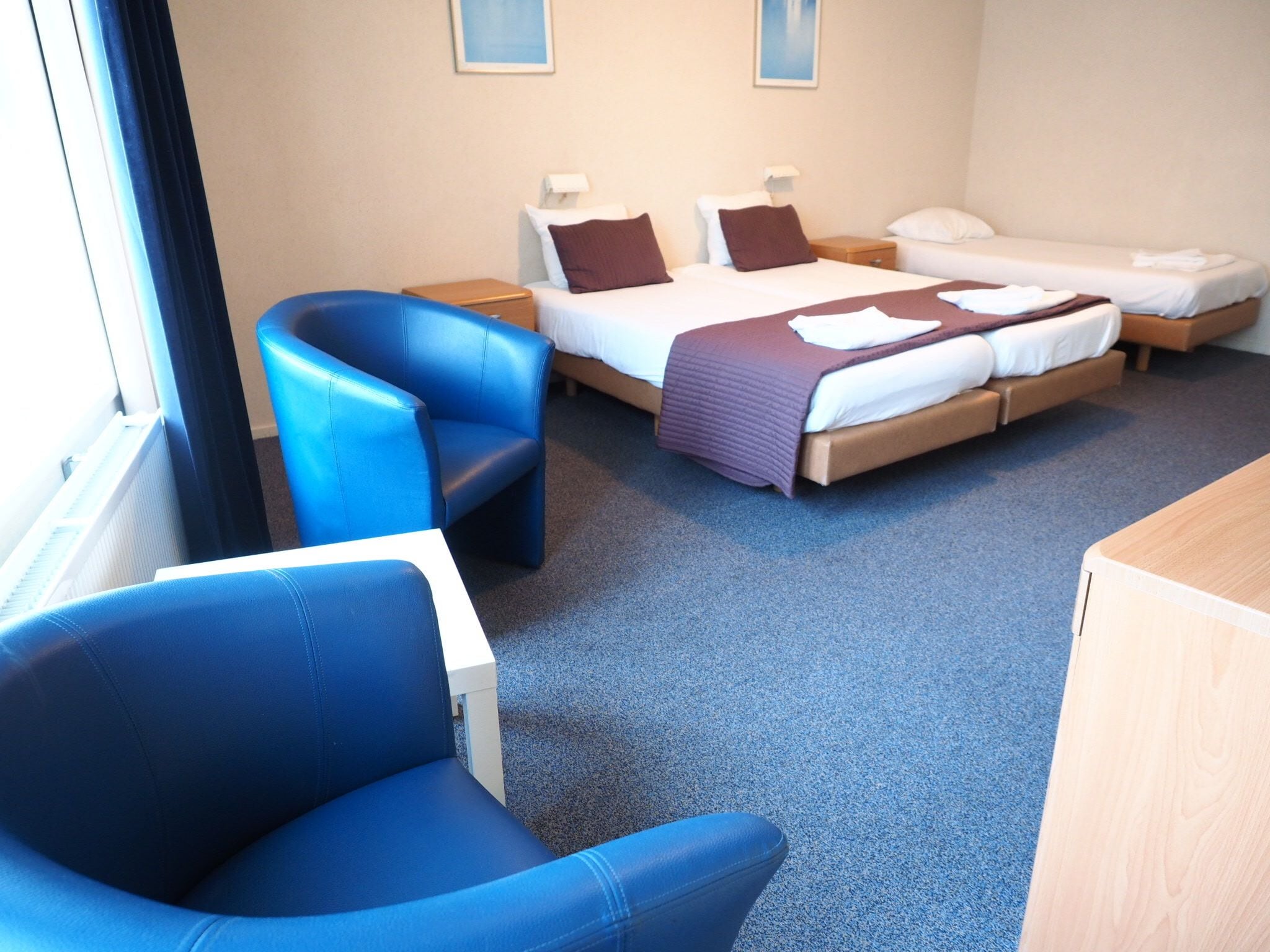 Una habitación de hotel con una cama, dos mesitas de noche, un sillón azul, un escritorio de madera y una ventana.