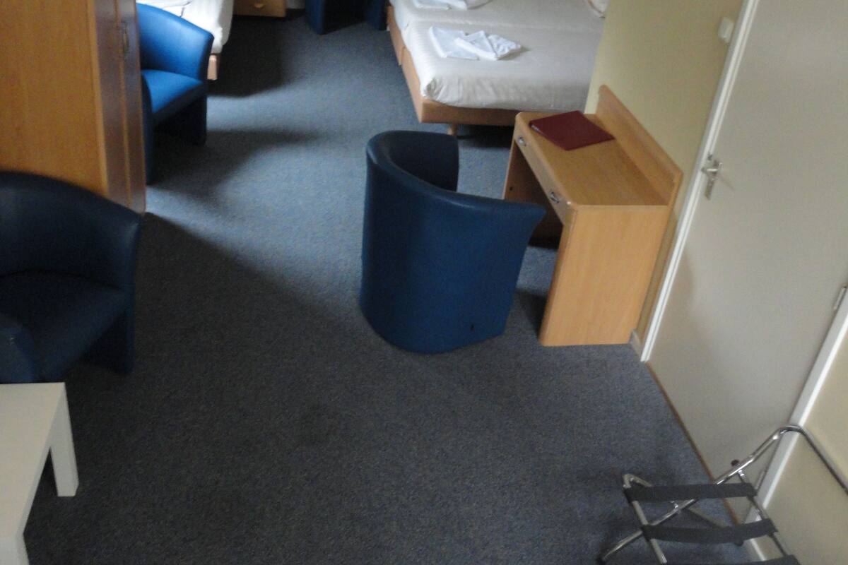 A compact hotel room with a single bed, a desk, a chair, and a small trash bin.