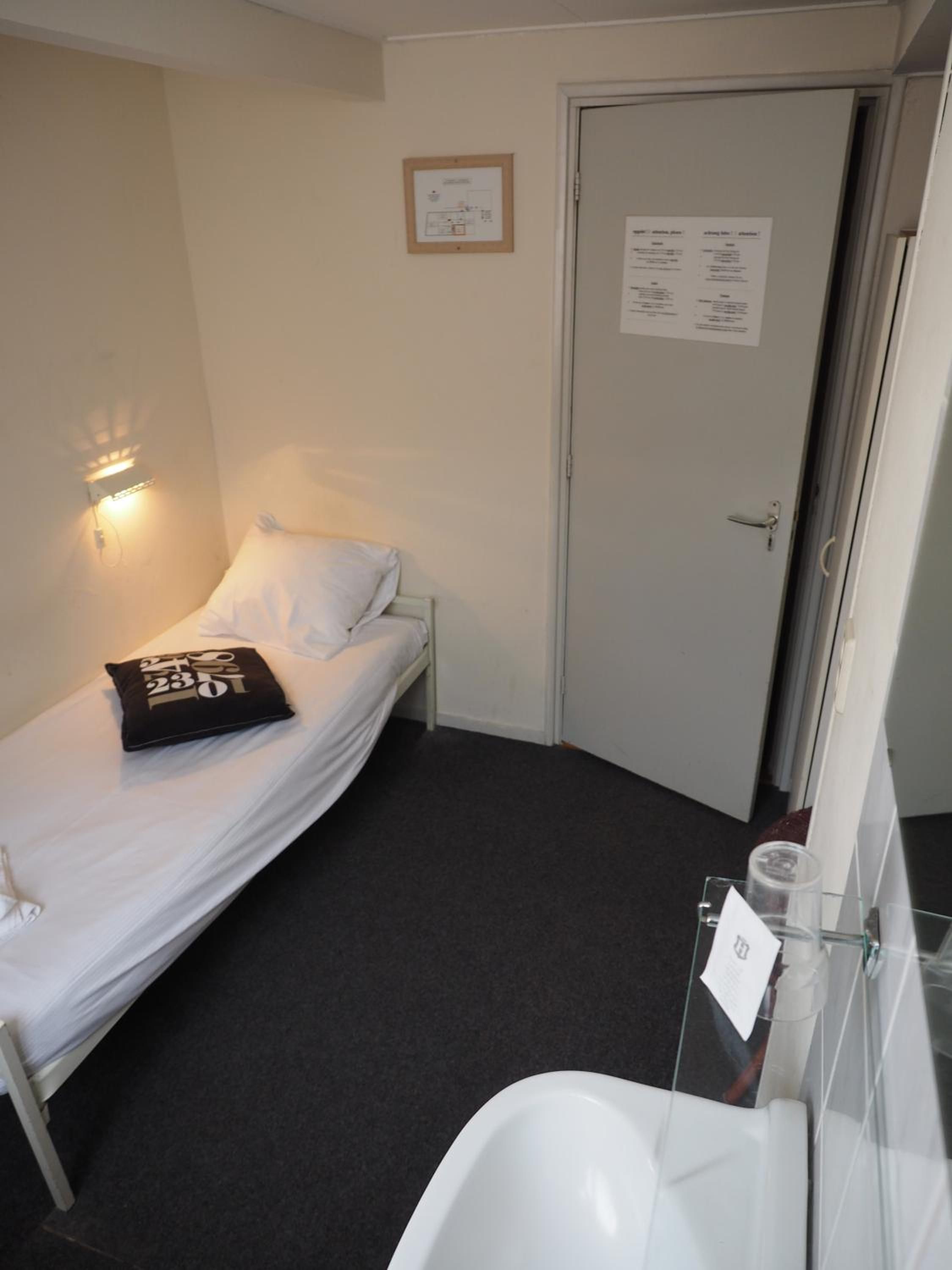 Una cama individual con un almohadón y una manta, una mesita de noche con un recipiente de vidrio, una lámpara de pared, una puerta con un aviso, y un baño con un lavamanos blanco.