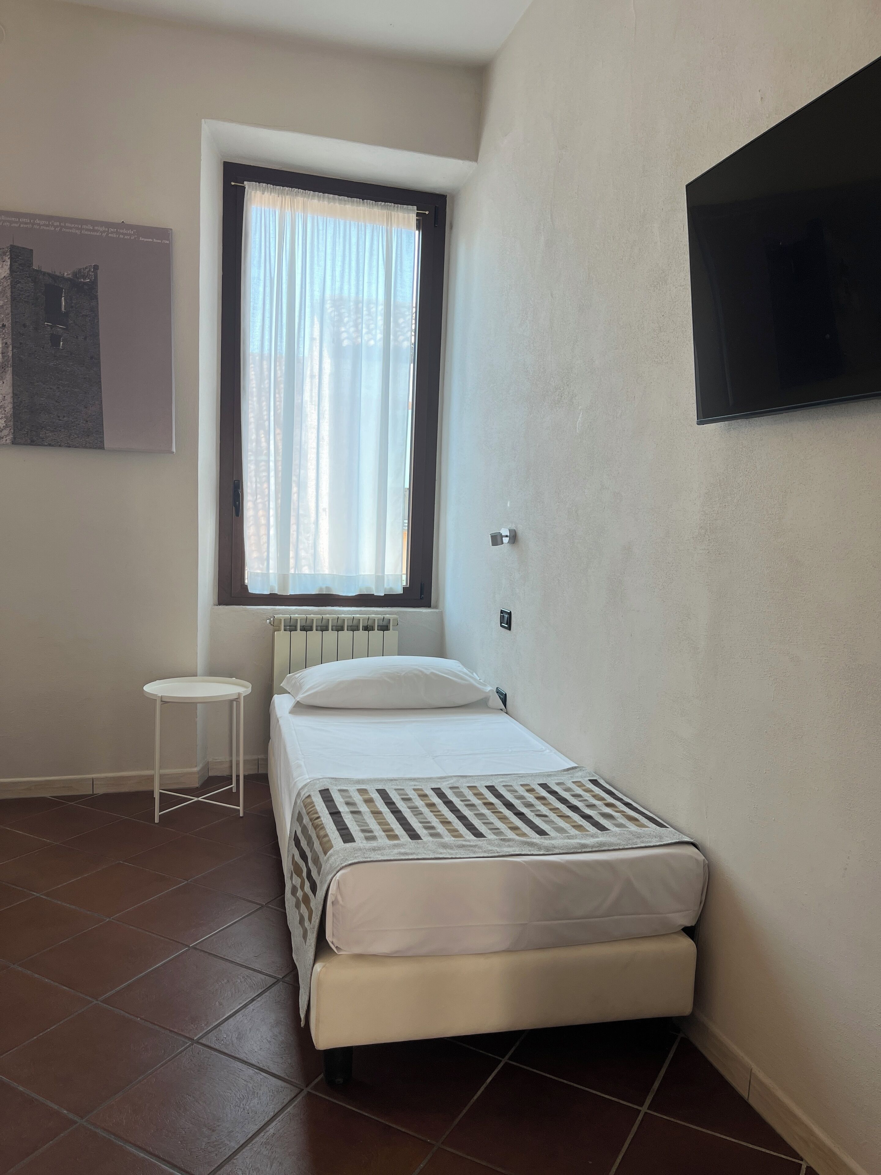 Una habitación pequeña y ordenada con una cama individual, una mesita, un radiador, una ventana con cortinas y un televisor montado.