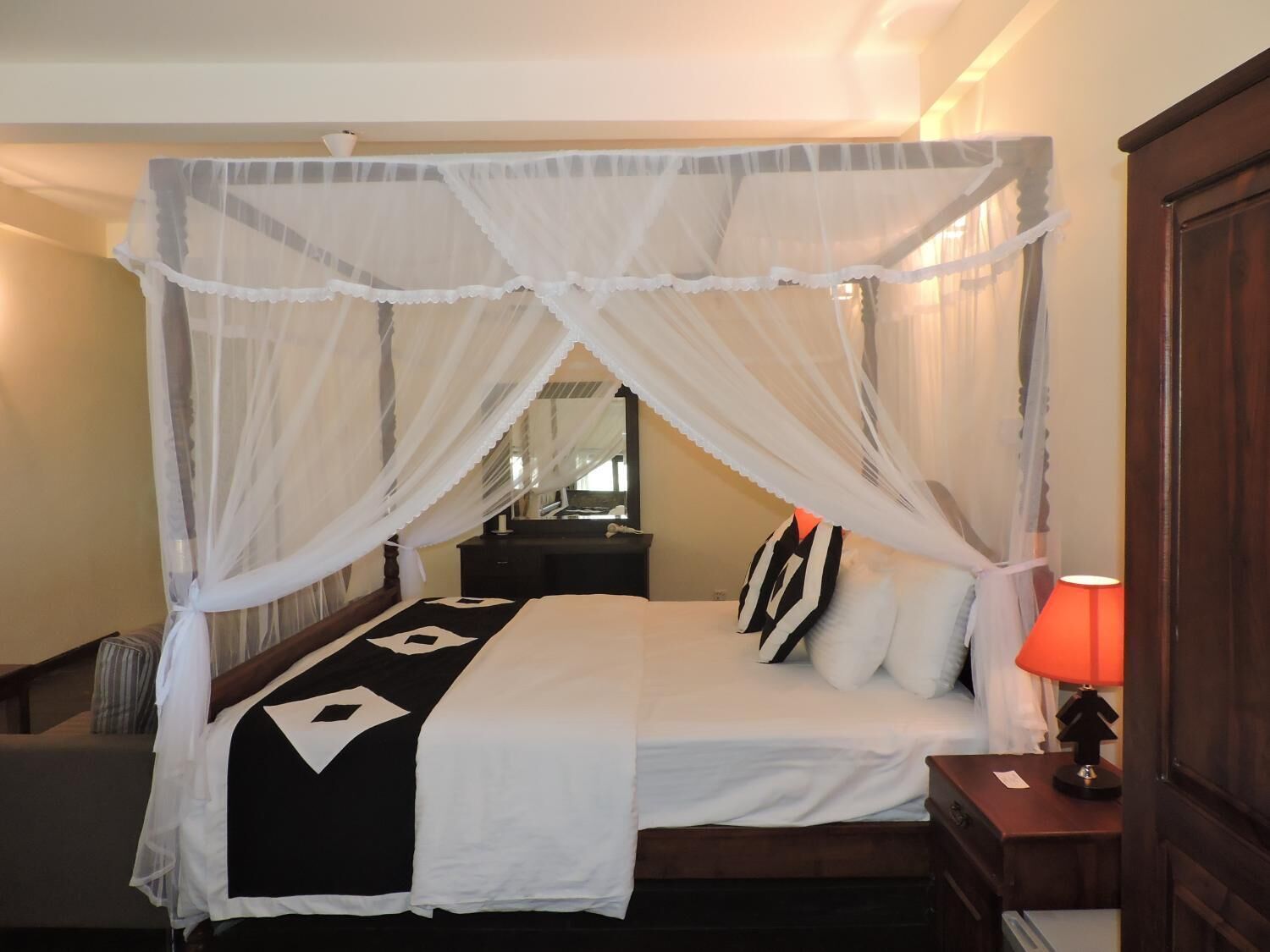 A hotel room with a four-poster canopy bed, a bedside table with a lamp, and a mirror on the wall.