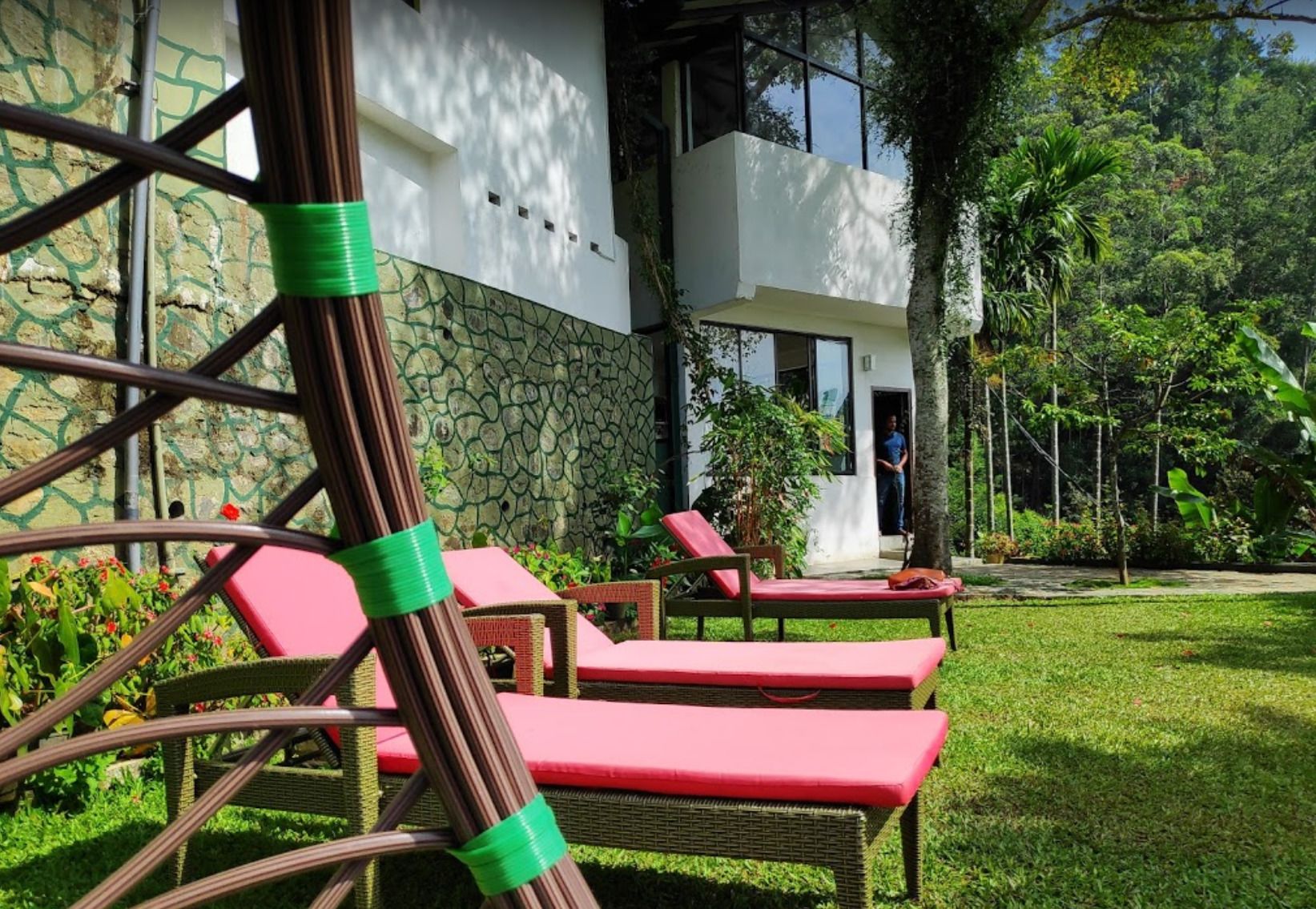 Un edificio moderno con facciata in pietra, ingresso con porta in vetro e un giardino curato con sedie in vimini e cuscini rossi.