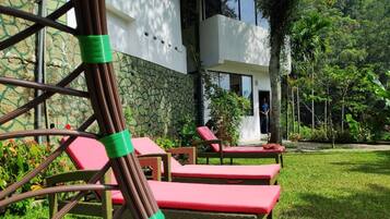 Un edificio moderno con facciata in pietra, ingresso con porta in vetro e un giardino curato con sedie in vimini e cuscini rossi.