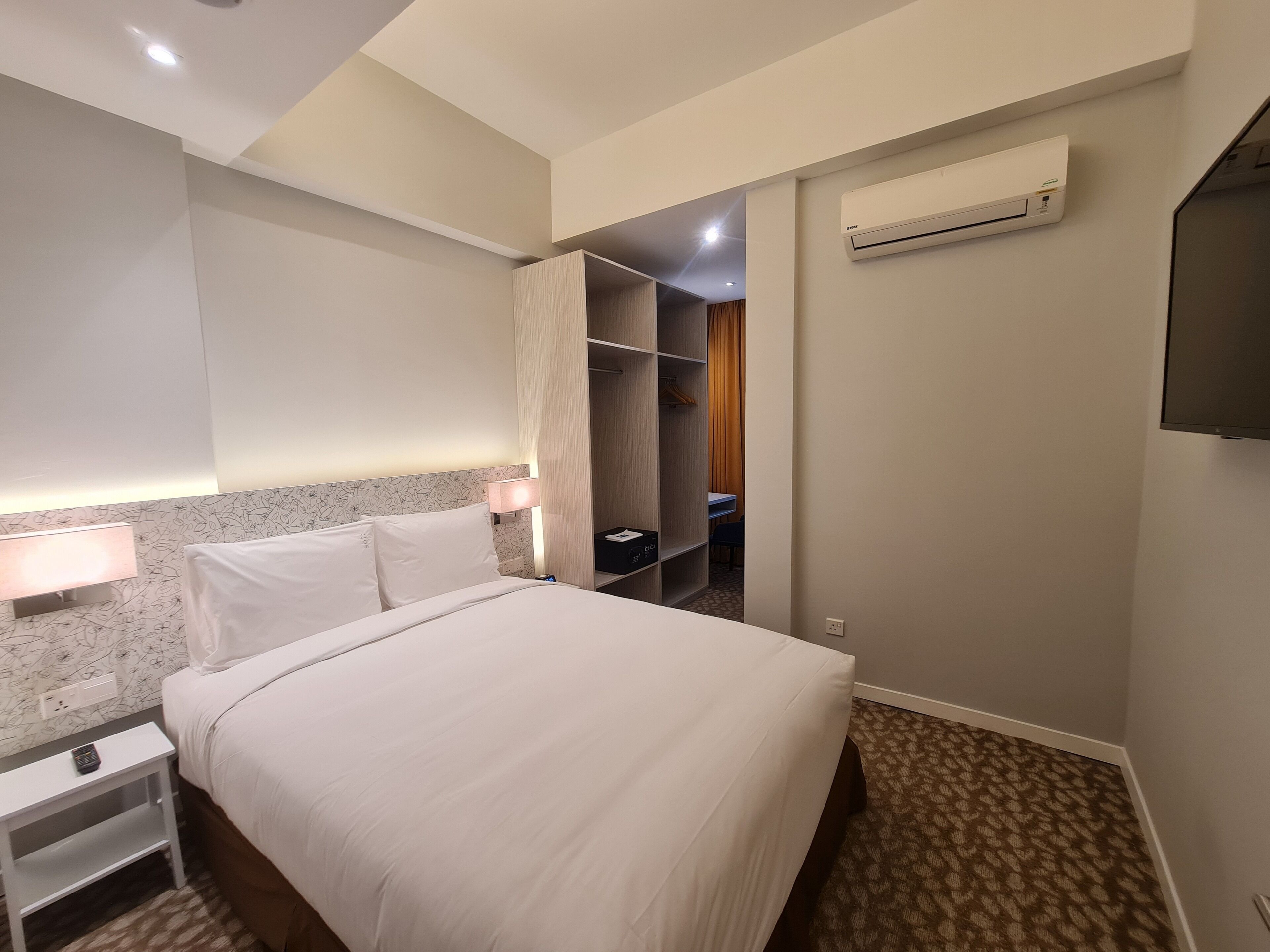 A hotel room with a bed, bedside table, lamp, wardrobe, and an air conditioning unit.