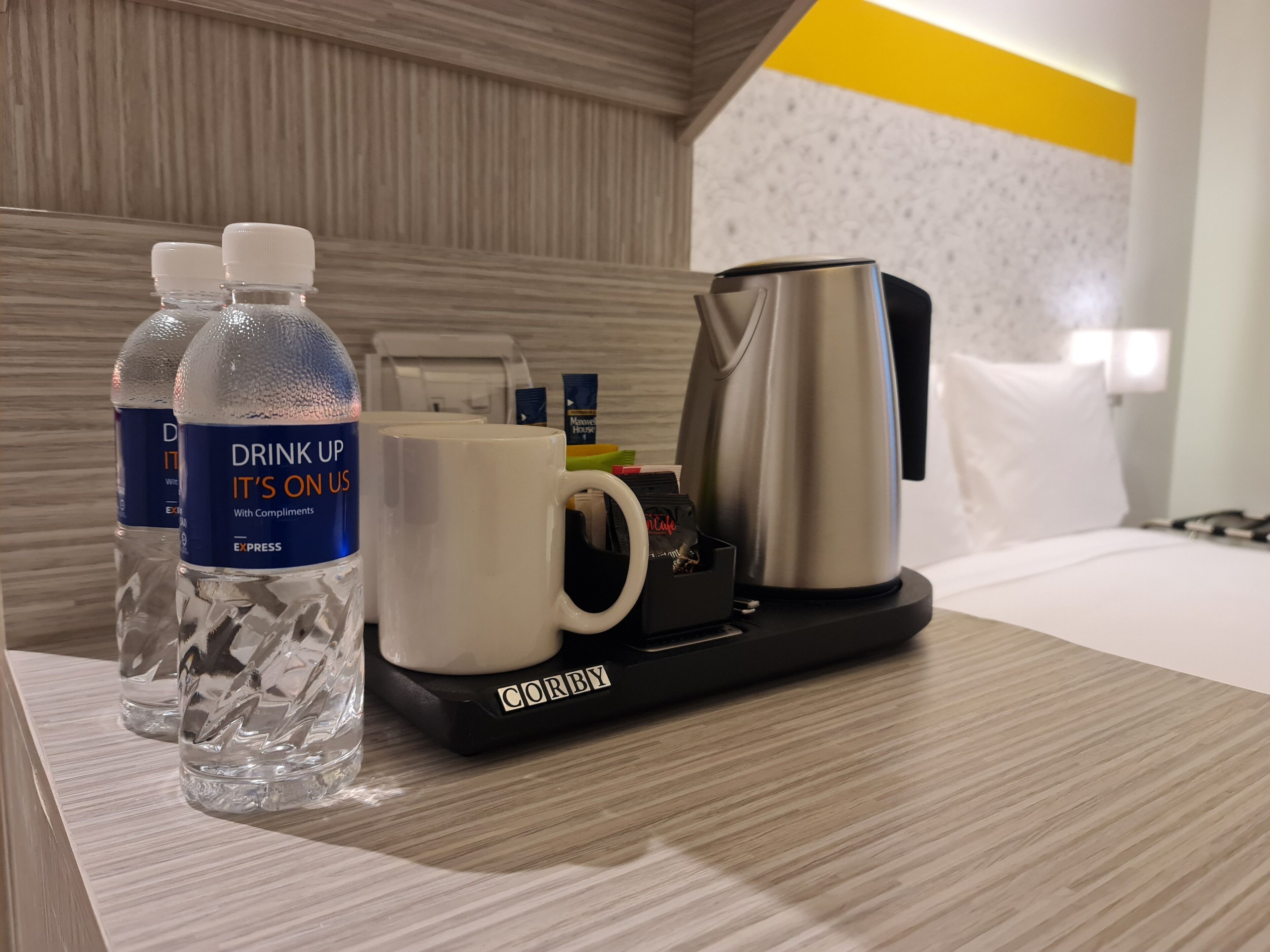 A hotel room with a wooden floor, a bed with white bedding, a black tray with a coffee maker, two water bottles, and a white mug.